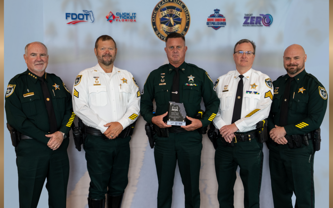Five uniformed law enforcement officers stand indoors, with one holding a clear award Behind them is a backdrop featuring Florida safety campaign logos and a palm tree image