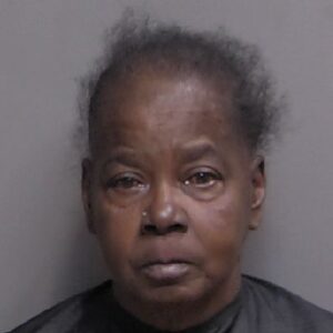 An older woman with short, thin hair is looking straight at the camera against a neutral gray background She has a serious expression and is wearing a dark top