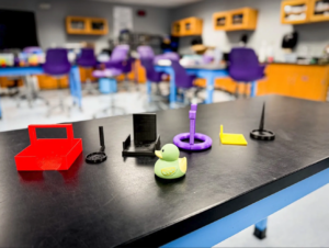 A lab table displays colorful D printed objects and a small rubber duck, with a classroom featuring purple chairs and lab equipment blurred in the background