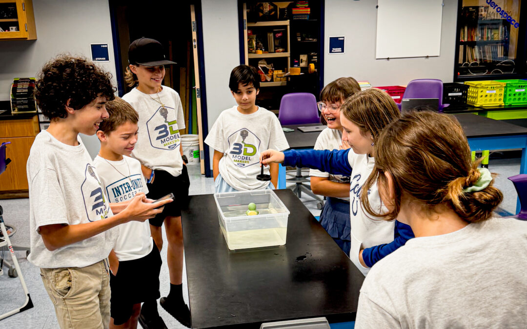 Seven kids in matching camp shirts gather around a table, watching an experiment with floating objects in a clear bin of water, smiling and engaged in a classroom setting