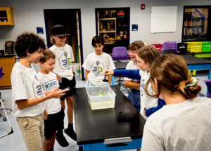 Seven kids in matching camp shirts gather around a table, watching an experiment with floating objects in a clear bin of water, smiling and engaged in a classroom setting