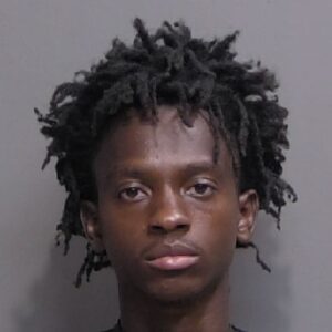 A mugshot of a young man with dreadlocks wearing a black shirt, photographed against a gray background