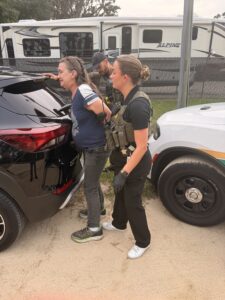 A woman in handcuffs is being searched by an officer next to a black car, while another officer stands nearby There are police vehicles and an RV in the background