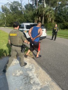 A sheriff escorts a handcuffed man in a blue tank top and red shorts toward a police SUV while another officer stands nearby on a sunny, rural road