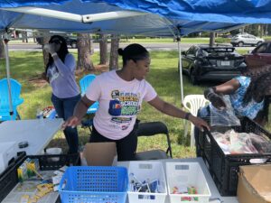 Three women organize school supplies under a blue tent outdoors One woman arranges items in bins, another drinks from a cup, and the third handles plastic bags Empty chairs and parked cars are visible in the background