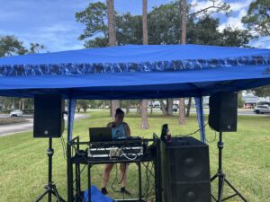 A person stands under a blue canopy, operating DJ equipment and a laptop on a table with speakers set up on grass Trees and cars are visible in the background on a partly cloudy day