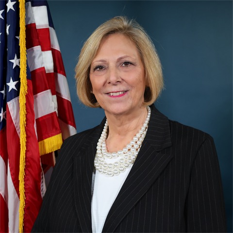 A woman with short blonde hair in a black pinstripe blazer and pearl necklace stands in front of an American flag, smiling at the camera with a blue background behind her