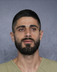 A man with short dark hair, a full beard, and a beige shirt stands against a gray background, looking slightly to the side with a neutral expression