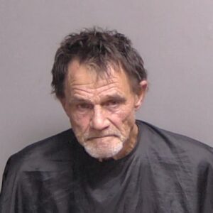 An older man with short, messy hair and a gray beard is wearing a dark cape against a plain gray background, looking downward with a serious expression An older man with short, messy hair and a gray beard is wearing a dark cape against a plain gray background, looking downward with a serious expression