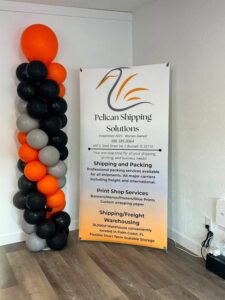A tall balloon column in orange, black, gray, and white stands beside a Pelican Shipping Solutions sign, detailing shipping and printing services in a bright, clean room