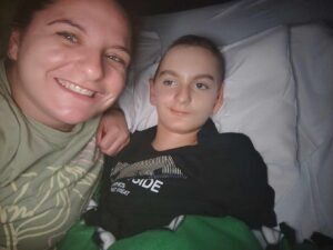 A smiling woman takes a selfie next to a boy lying in bed with a pillow behind his head, covered by a green and black blanket