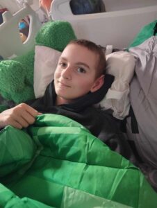 A young person with short hair lies in bed, covered with a green blanket, surrounded by pillows, looking at the camera with a calm expression