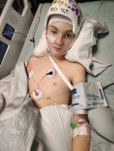 A young person lies in a hospital bed wearing a protective helmet, with medical monitors, a blood pressure cuff, and an IV line attached, looking at the camera