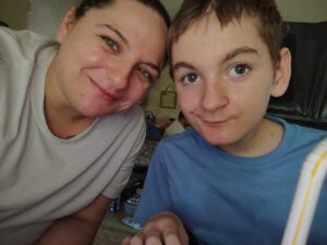 A woman and a boy are smiling at the camera indoors The woman is wearing a light gray shirt, and the boy is in a blue shirt They appear to be close together, with a casual, relaxed atmosphere
