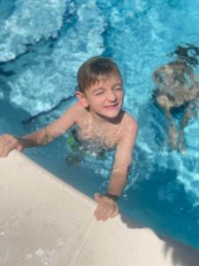 A young boy with short hair smiles and squints in the sunlight while holding onto the edge of a swimming pool, partially submerged in the clear blue water