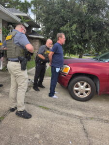 Two sheriff officers handcuff a man in a blue shirt and socks beside a red SUV parked in a driveway, near a brick house and trees