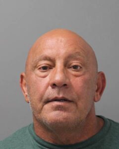 A middle aged bald man with tanned skin wearing a green shirt, looking directly at the camera with a neutral expression against a plain gray background