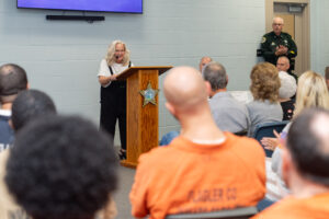 An older woman speaks at a podium with a sheriff’s star emblem, facing an audience that includes people in orange uniforms A uniformed officer stands nearby against a light blue wall