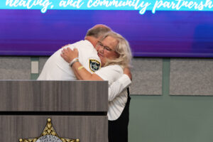 A man in a sheriff’s uniform hugs a smiling woman at a podium A blue screen in the background displays partially visible text A man in a sheriff’s uniform hugs a smiling woman at a podium A blue screen in the background displays partially visible text