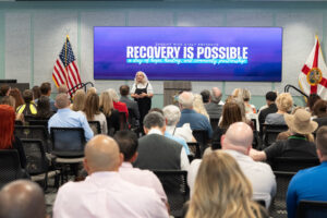 A woman speaks to an audience in a conference room Behind her, a large screen displays “Recovery Is Possible” next to American and Florida flags The audience sits facing her, listening attentively A woman speaks to an audience in a conference room Behind her, a large screen displays “Recovery Is Possible” next to American and Florida flags The audience sits facing her, listening attentively