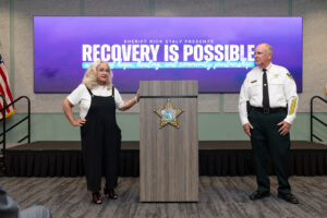 A woman speaks at a podium with a sheriff standing nearby The screen behind them reads, “RECOVERY IS POSSIBLE” and mentions hope, healing, and community partnerships An American flag is visible on the left A woman speaks at a podium with a sheriff standing nearby The screen behind them reads, “RECOVERY IS POSSIBLE” and mentions hope, healing, and community partnerships An American flag is visible on the left