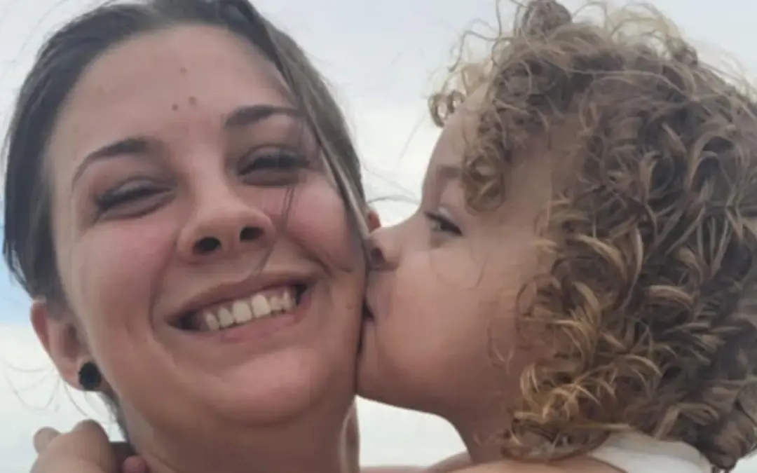 A young child with curly hair kisses a smiling woman on the cheek The woman is holding the child, and both appear happy, outdoors, with a cloudy sky in the background