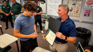 A man hands a certificate to a smiling student in a classroom, while others in the background look on and smile Classroom posters and desks are visible