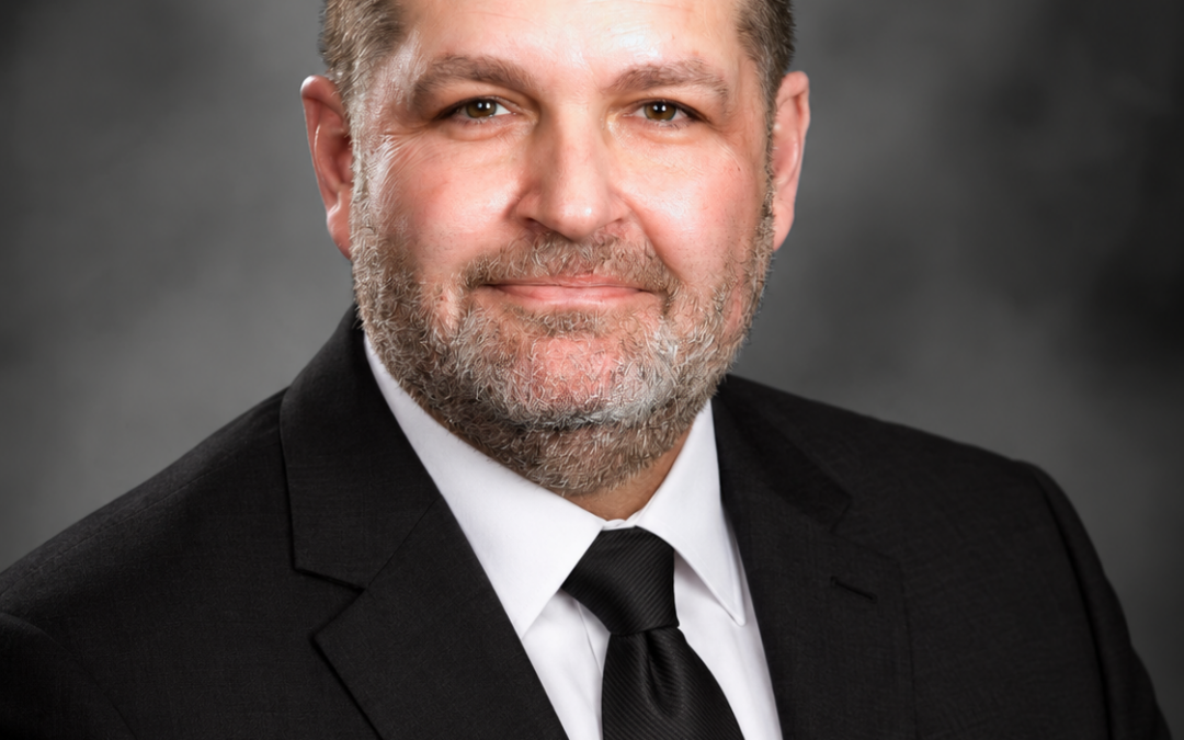 A man with short dark hair and a trimmed beard, wearing a black suit, white shirt, and black tie, poses for a professional portrait against a dark, blurred background