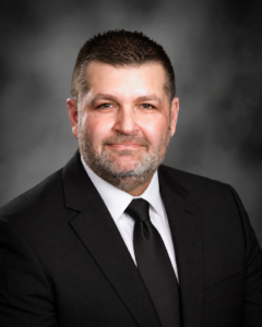 A man with short dark hair and a trimmed beard, wearing a black suit, white shirt, and black tie, poses for a professional portrait against a dark, blurred background A man with short dark hair and a trimmed beard, wearing a black suit, white shirt, and black tie, poses for a professional portrait against a dark, blurred background