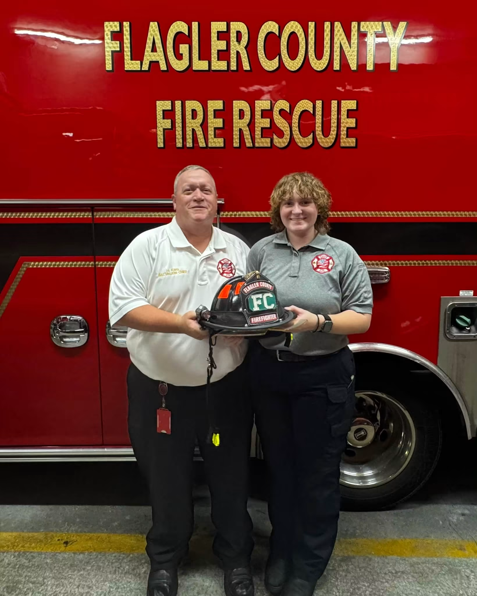Two firefighters stand in front of a red Flagler County Fire Rescue truck, smiling and holding a black fire helmet together Both wear uniforms, and the truck’s large yellow lettering is visible behind them