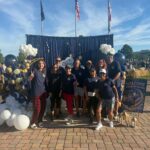 A group of people pose in front of a balloon arch and backdrop with American flags in the background at an outdoor event on a sunny day
