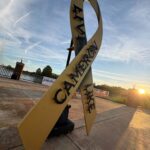A large gold ribbon sculpture stands outdoors near a fence at sunrise The words “LIVE LIKE CAMERON” are displayed along the ribbon