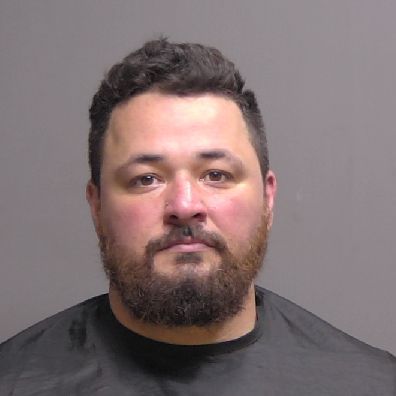 A man with short dark hair and a beard is facing the camera against a plain gray background, wearing a dark shirt or drape