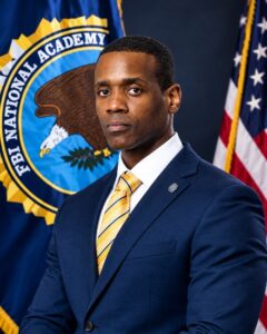 A man in a blue suit stands in front of an FBI National Academy flag and a U S flag, looking serious, with a yellow striped tie and a pin on his lapel