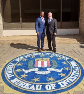 Two men in suits stand smiling on an outdoor FBI seal, located in front of a building with large windows The seal reads “Department of Justice Federal Bureau of Investigation ”