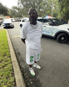 A man in white clothes stands on a street near police cars with flashing lights He has sunglasses, a gold chain, and his hands behind his back Trees and grass are visible in the background