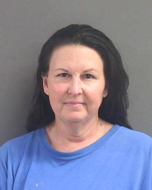 A headshot of a middle aged woman with dark hair wearing a blue crew neck shirt against a gray background
