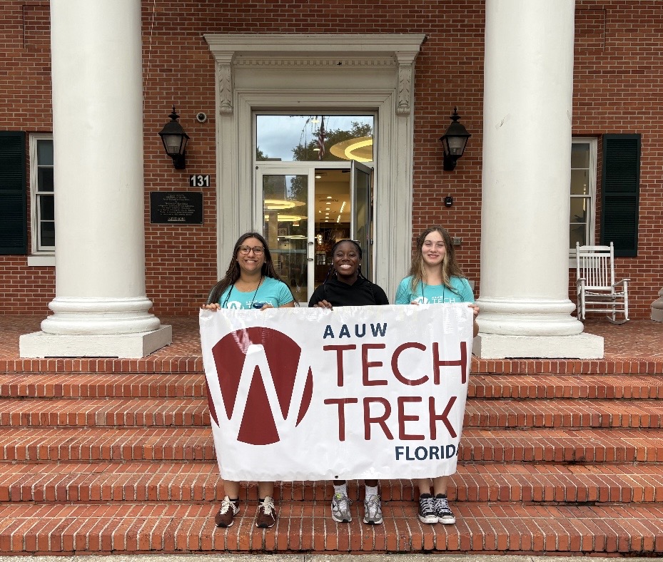 Three young women stand on brick steps in front of a building holding a banner that reads, “AAUW Tech Trek Florida ” Three young women stand on brick steps in front of a building holding a banner that reads, “AAUW Tech Trek Florida ”