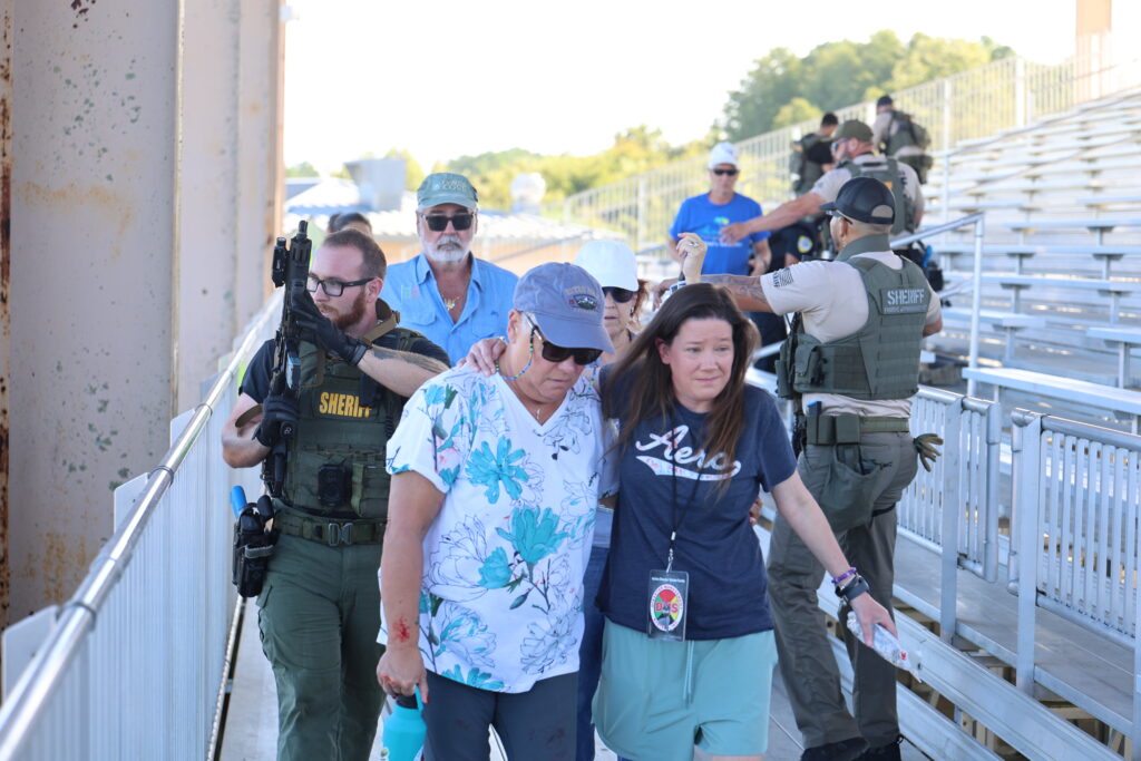 Two women walk together in front of bleachers, one with a bandaged arm Several sheriff officers and people in blue shirts are present, creating a tense scene