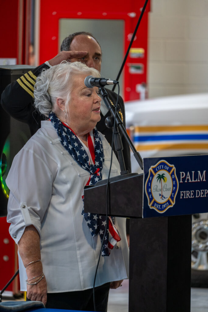 A woman with white hair wearing a star spangled scarf speaks at a podium with the Palm Coast Fire Department logo, as a uniformed man stands behind her saluting A woman with white hair wearing a star spangled scarf speaks at a podium with the Palm Coast Fire Department logo, as a uniformed man stands behind her saluting