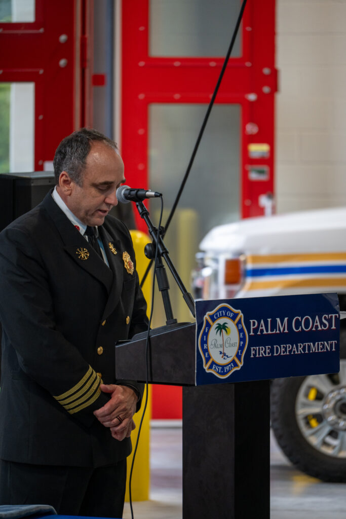 A fire chief in a formal uniform stands at a podium with the Palm Coast Fire Department seal, speaking into a microphone A fire chief in a formal uniform stands at a podium with the Palm Coast Fire Department seal, speaking into a microphone