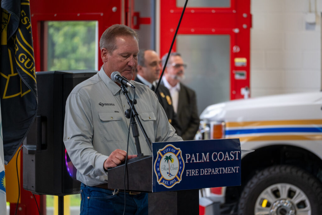 A man in a light colored long sleeve shirt speaks at a podium labeled “City of Palm Coast Fire Department ” A man in a light colored long sleeve shirt speaks at a podium labeled “City of Palm Coast Fire Department ”