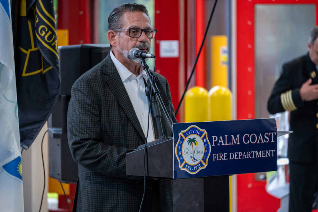 A man with glasses and a goatee speaks at a podium marked with the City of Palm Coast Fire Department logo A man with glasses and a goatee speaks at a podium marked with the City of Palm Coast Fire Department logo
