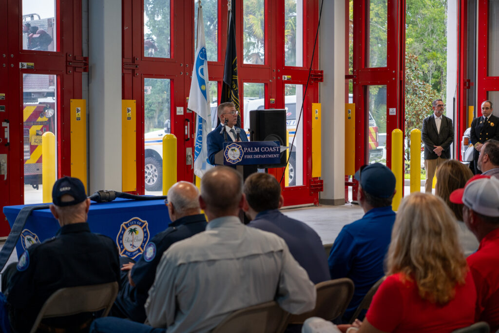 A man in a suit speaks at a Palm Coast Fire Department podium in front of a seated audience of firefighters and staff A man in a suit speaks at a Palm Coast Fire Department podium in front of a seated audience of firefighters and staff