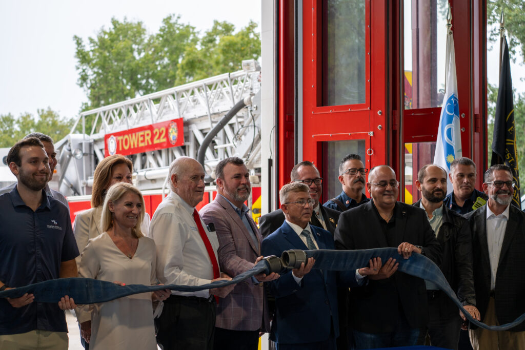 A group of people standing in front of a fire station participate in a ribbon cutting ceremony using a fire hose, with a fire truck labeled “Tower ” visible in the background A group of people standing in front of a fire station participate in a ribbon cutting ceremony using a fire hose, with a fire truck labeled “Tower ” visible in the background