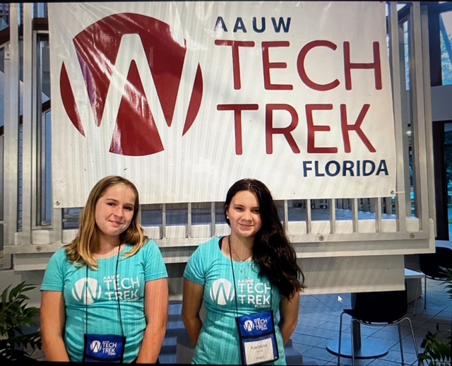 Two young women wearing matching teal AAUW Tech Trek t shirts and lanyards stand in front of an AAUW Tech Trek Florida banner Two young women wearing matching teal AAUW Tech Trek t shirts and lanyards stand in front of an AAUW Tech Trek Florida banner