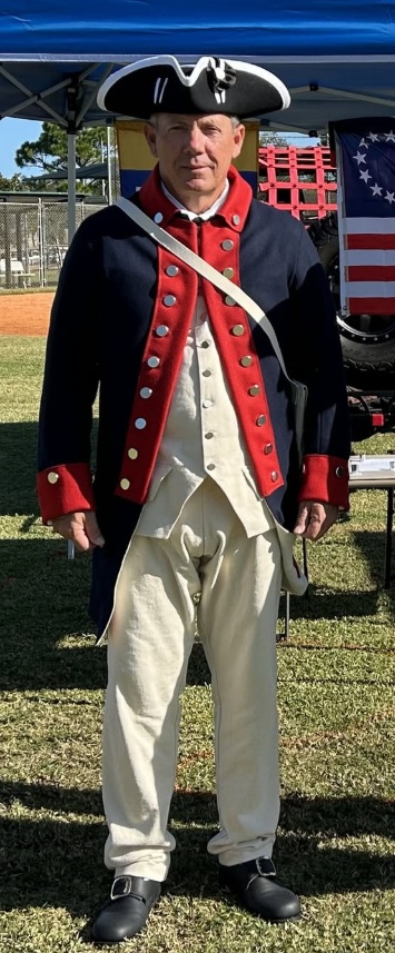 A man in an th century American Continental Army uniform including a blue coat with red facings, light colored breeches, and a tricorne hat