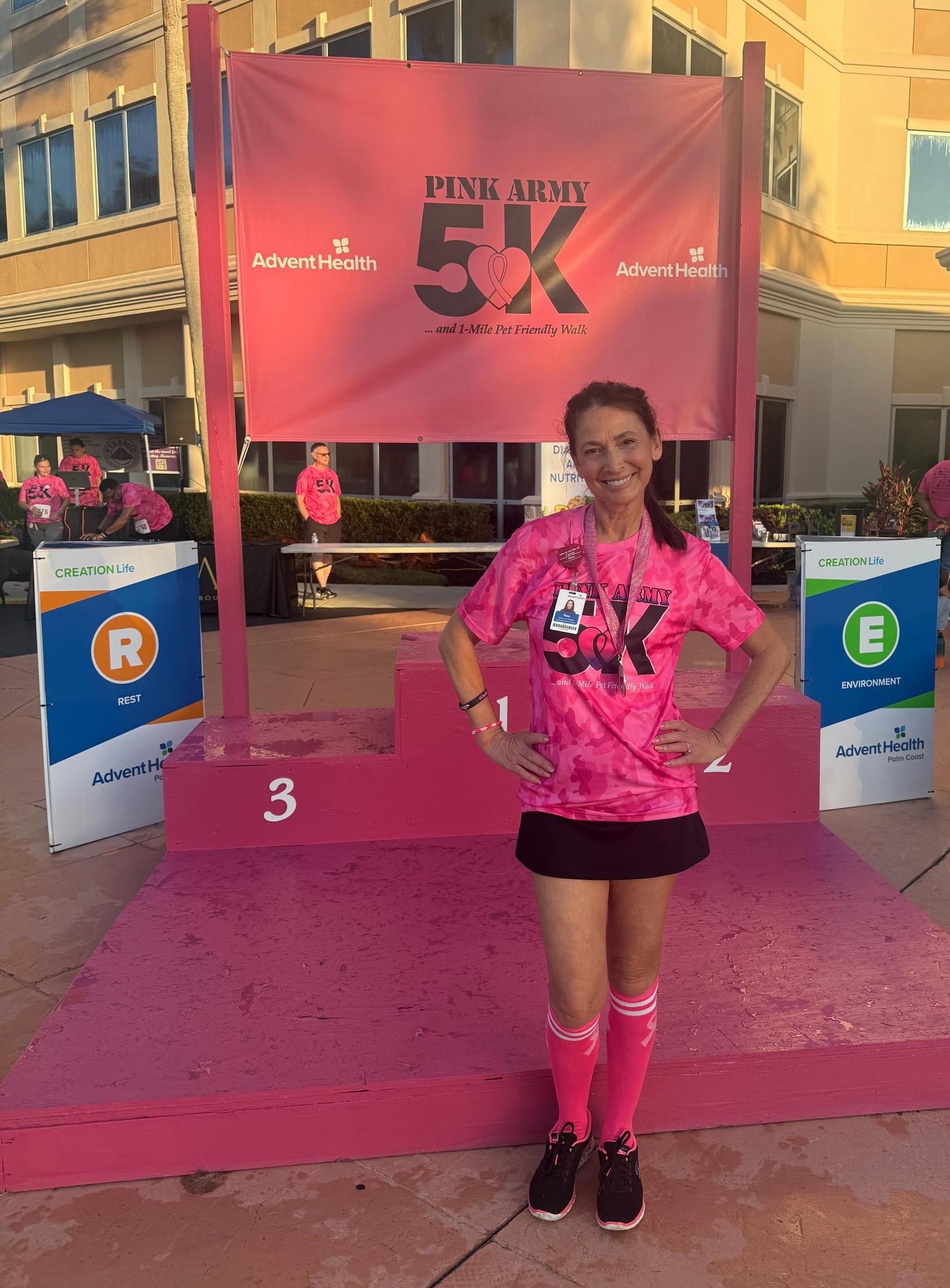 A woman wearing a pink race t shirt and finisher medal stands on a pink race podium in front of a Pink Army K sign A woman wearing a pink race t shirt and finisher medal stands on a pink race podium in front of a Pink Army K sign