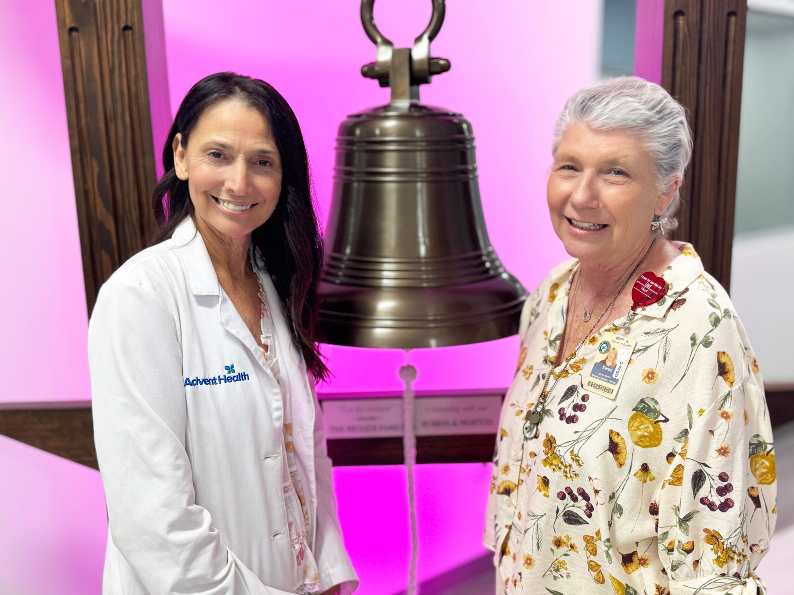 A healthcare provider in a white lab coat stands next to a woman wearing a floral blouse in front of a commemorative bell