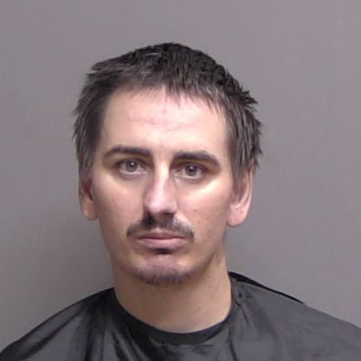Mugshot of a man with short brown hair and a thin mustache, wearing a black shirt against a gray background